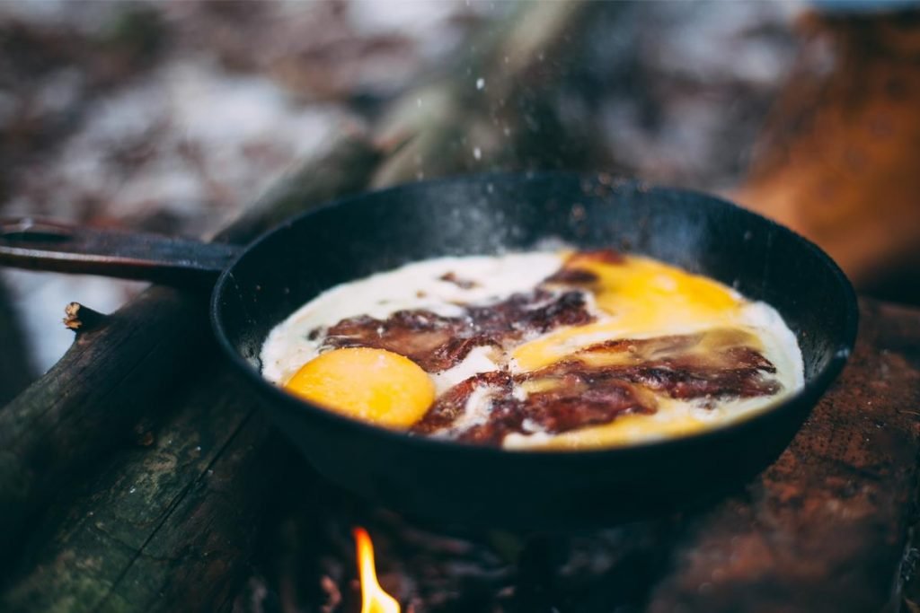 Egg and bacon cooking in a frying pan over campfire flame